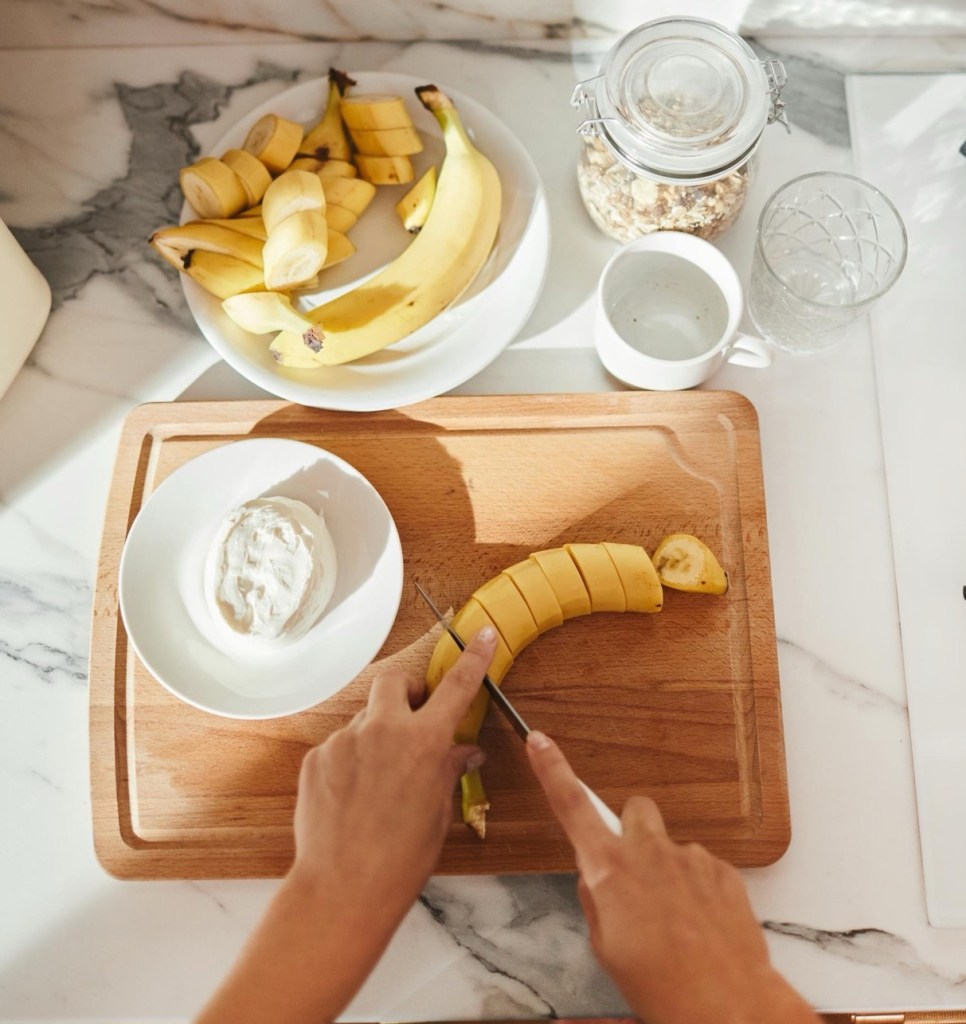 Hands slicing banana on a cutting board with Greek yogurt, bananas, and ginger ingredients prepared for a gut-soothing smoothie