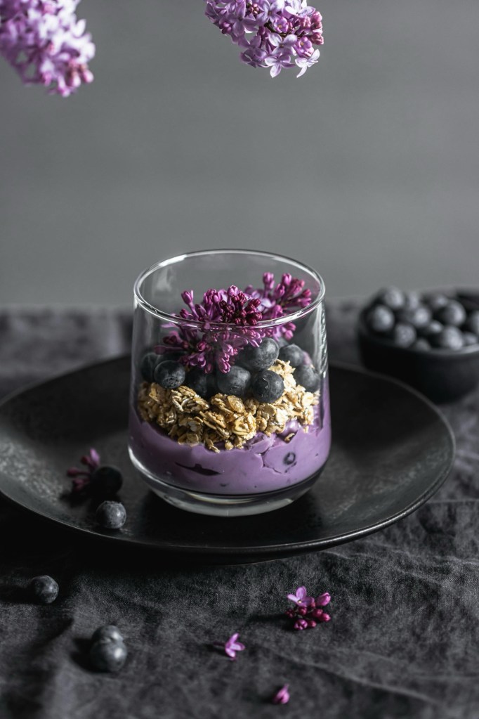 Glass cup filled with layers of blueberry yogurt, granola, and fresh blueberries topped with purple edible flowers