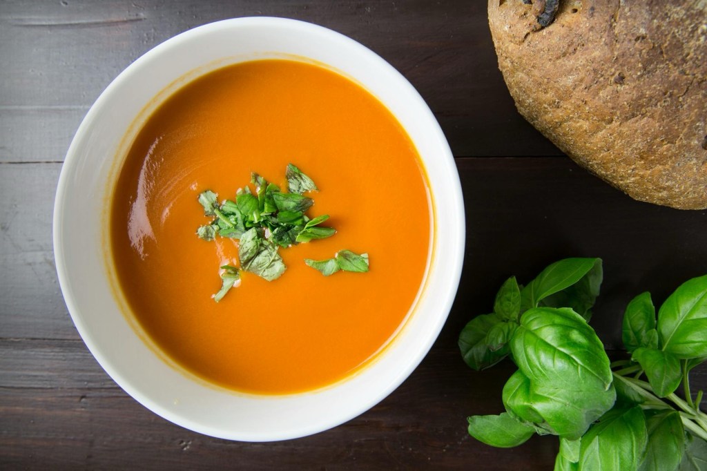 Bowl of bright orange lentil and vegetable soup topped with fresh herbs, next to a piece of whole grain bread and basil leaves